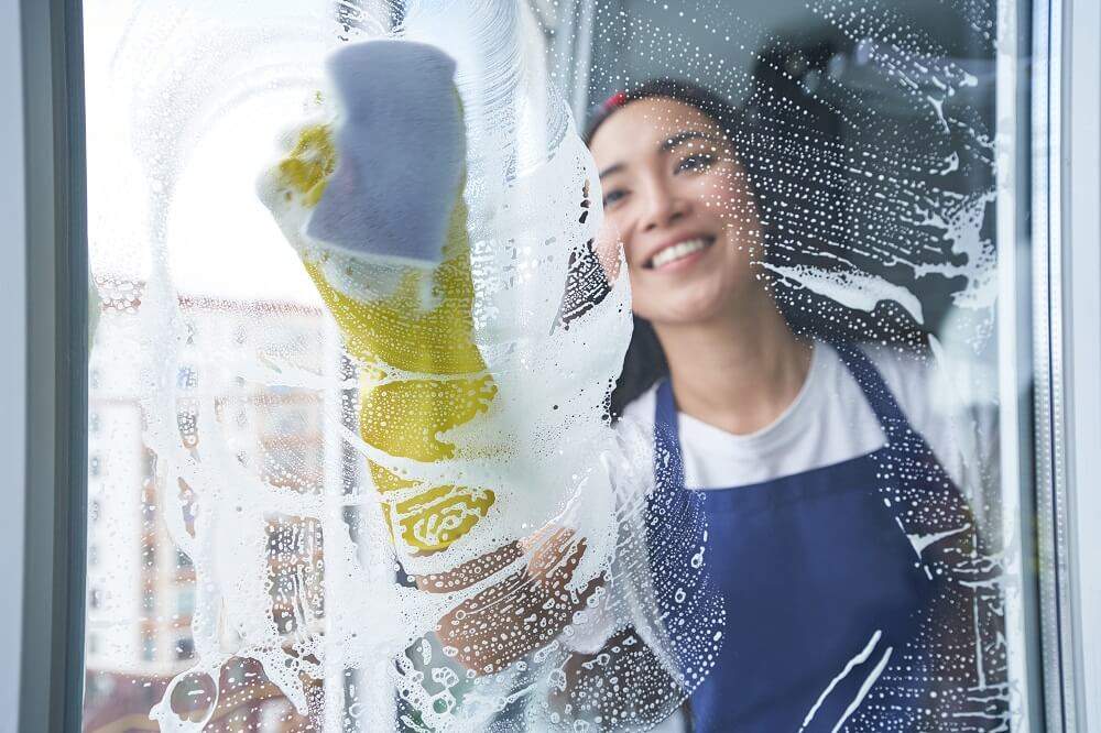 Cheerful young woman smiling while cleaning the window, glass surface using sponge. Housework and housekeeping, cleaning service concept. View through the glass